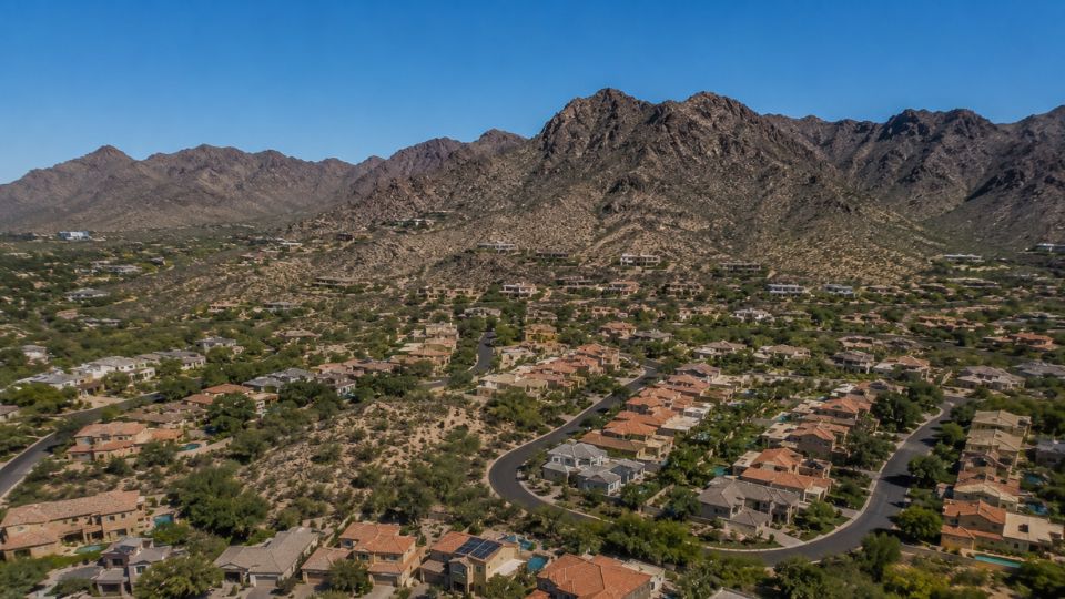 Aerial view of suburban desert neighborhood with tile-roof homes, winding streets and rugged mountain backdrop under blue sky
