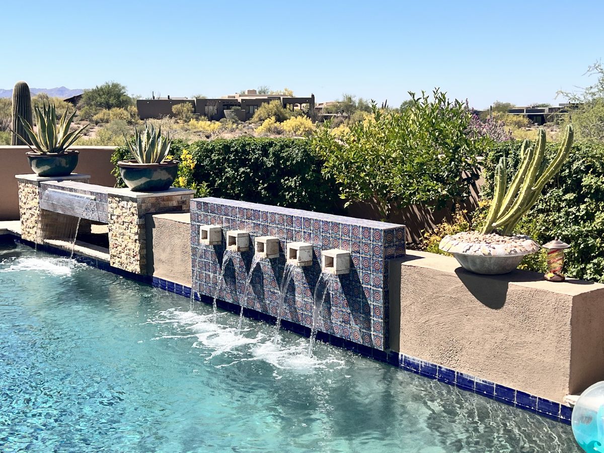 Resort-style backyard pool with patterned tiled wall fountains, cactus planters and desert landscape under blue sky