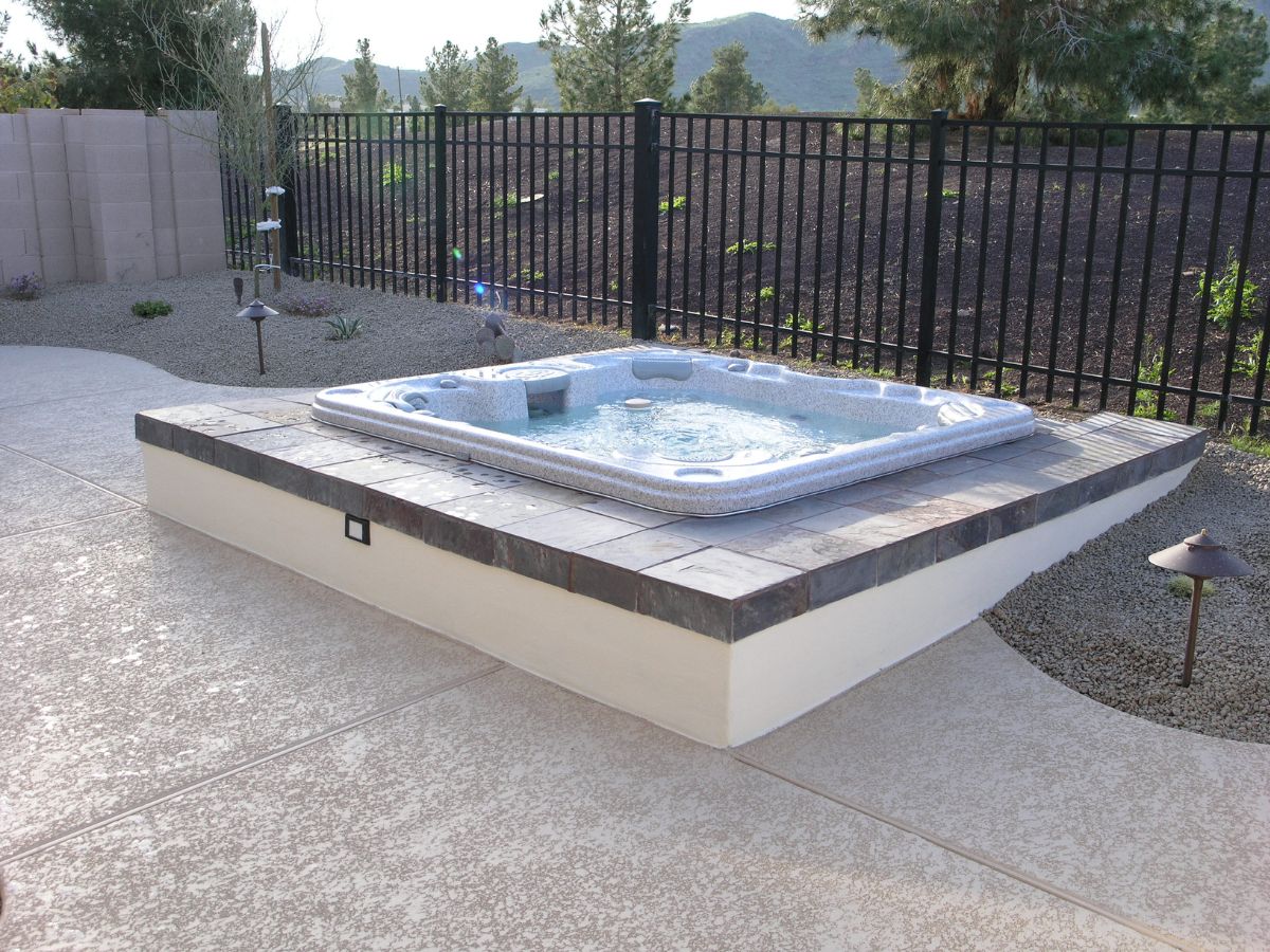 Outdoor backyard hot tub on raised tiled platform with patio, gravel landscaping and black metal fence with mountain view.