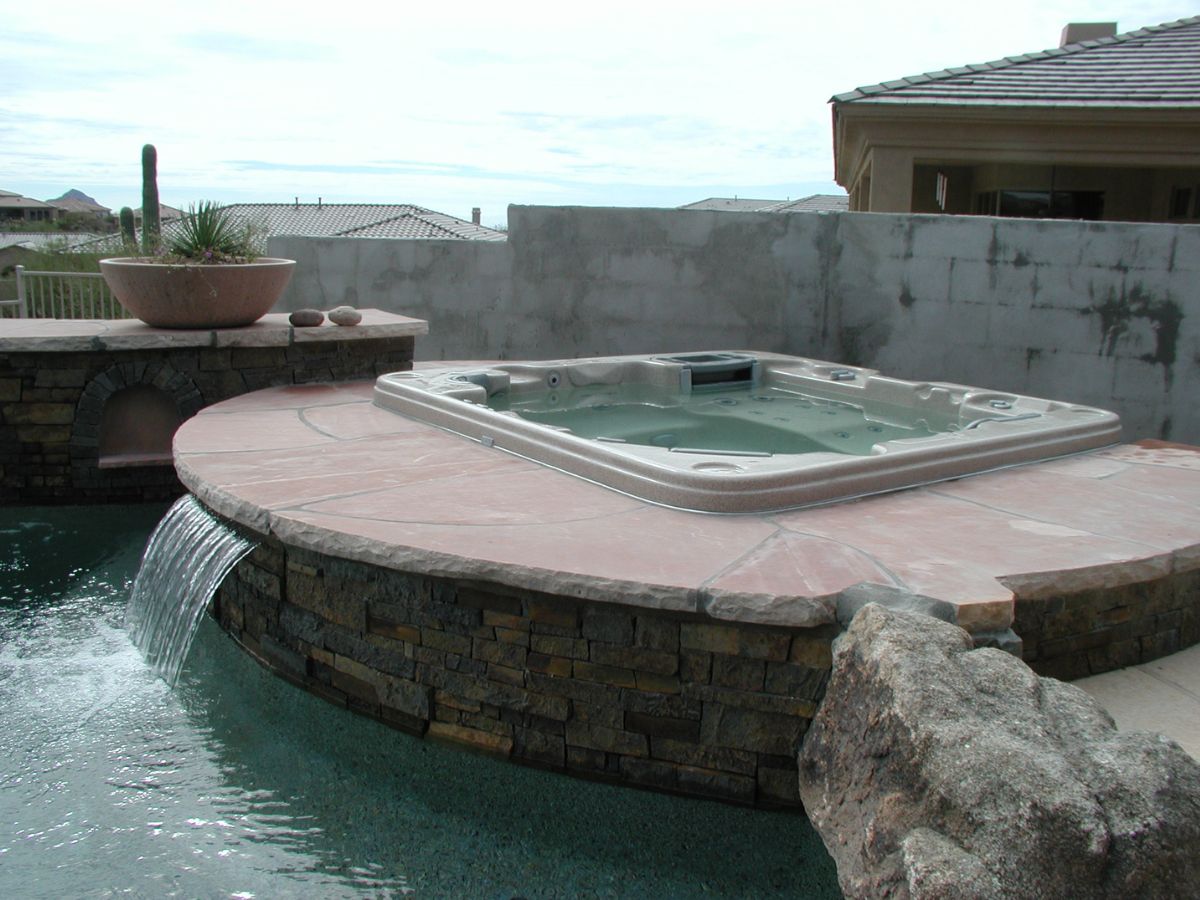 Luxury backyard hot tub with stone spillover waterfall flowing into swimming pool on desert-style patio