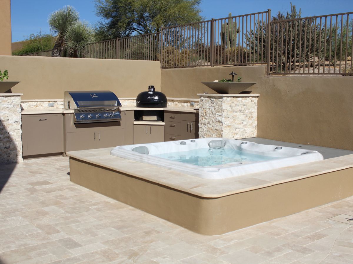 Luxury outdoor kitchen with stainless grill and kamado beside sunken hot tub on travertine patio in desert backyard