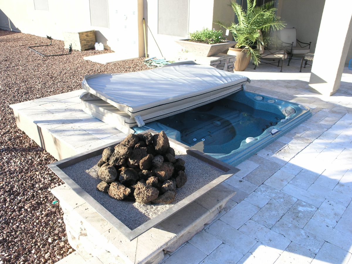 Built-in backyard hot tub with folded cover beside lava-rock fire pit on stone patio, potted plants and gravel landscaping.
