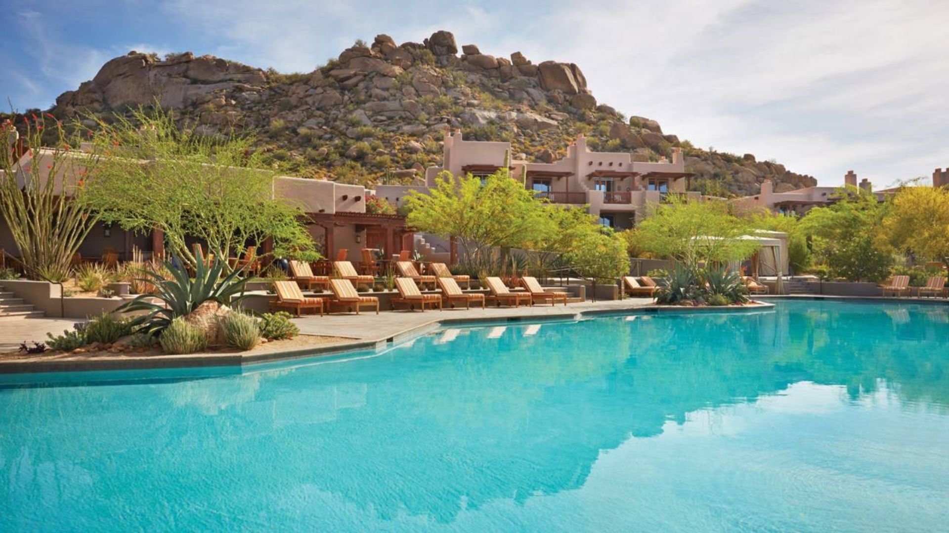 Luxury resort pool with blue water, wooden lounge chairs, desert plants and rocky mountain backdrop