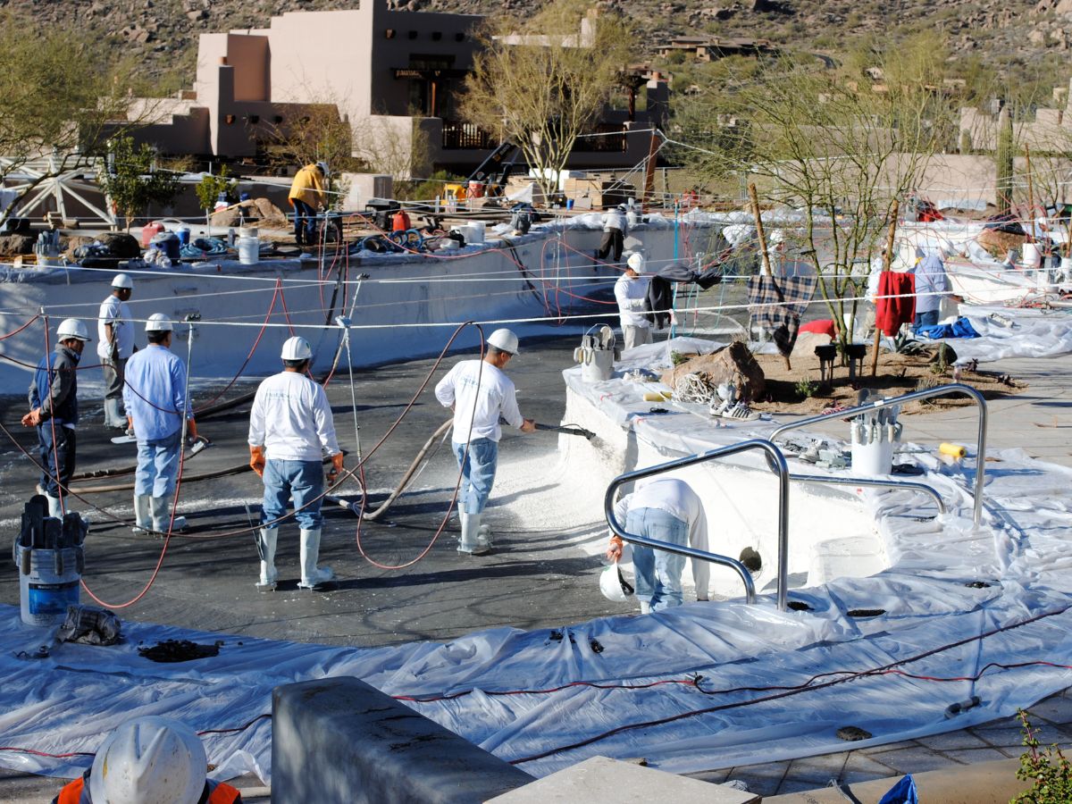 Workers in hard hats resurfacing large outdoor pool with white waterproof coating during commercial pool renovation