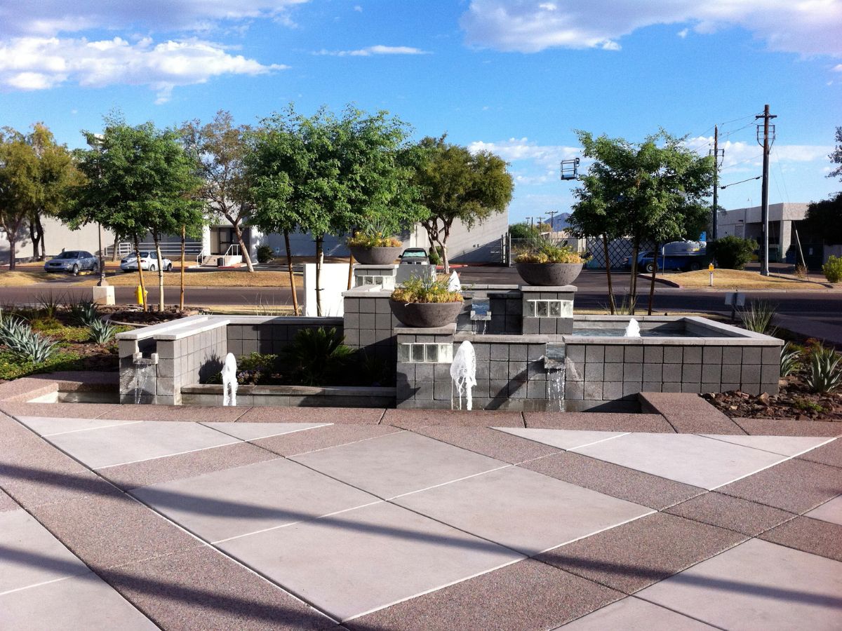 Tiered stone fountain in modern urban plaza with concrete planters, small trees and clear blue sky