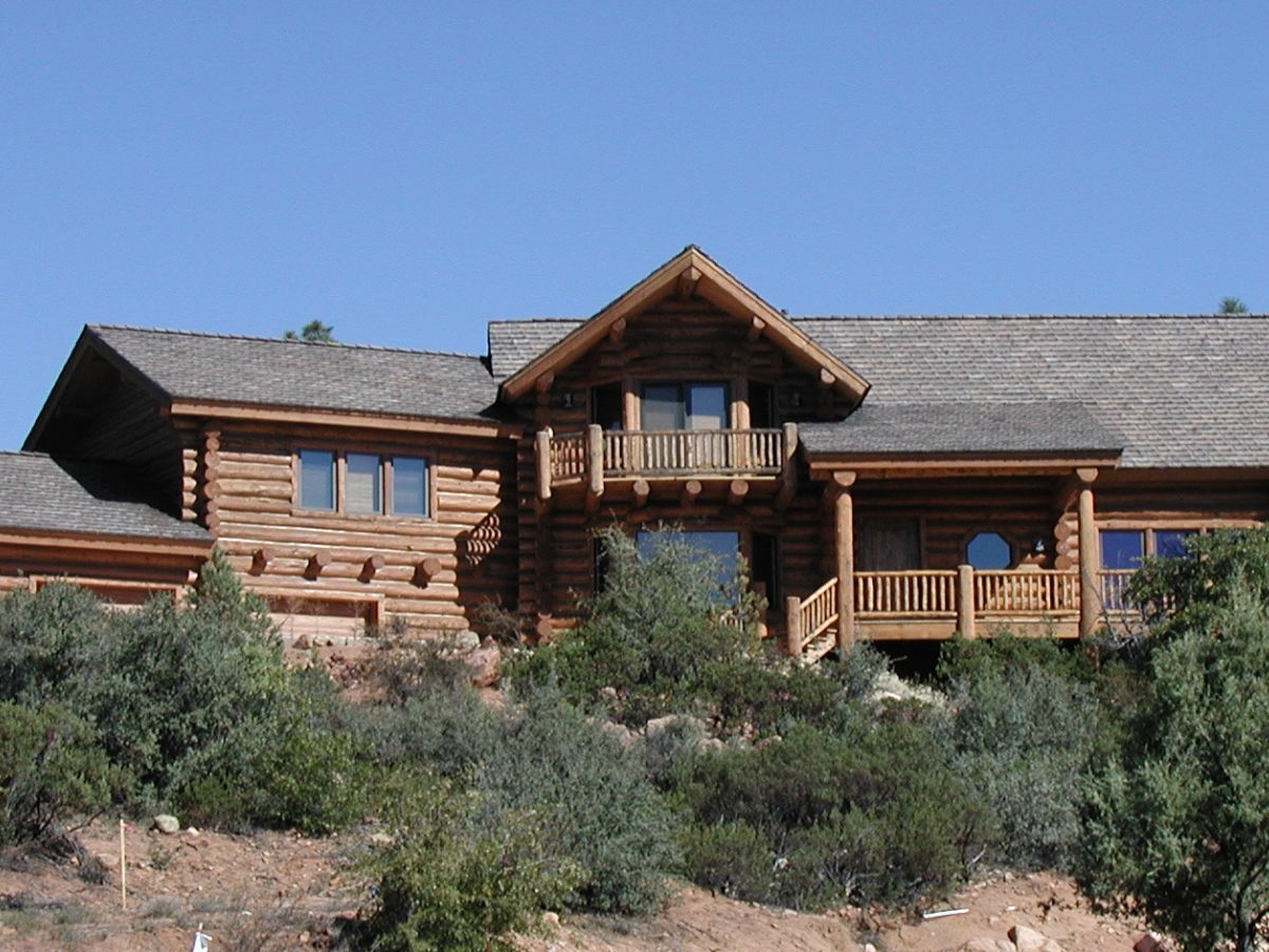 Rustic log cabin mountain home with wooden balcony and porch on hillside surrounded by shrubs under clear blue sky