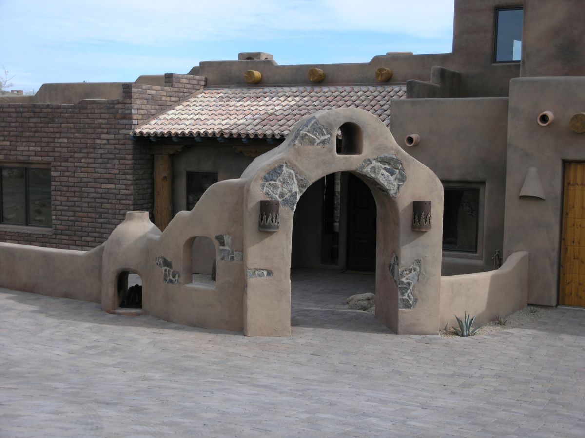 Southwestern adobe house entrance with arched stucco gateway, stone accents, tiled roof and paved courtyard