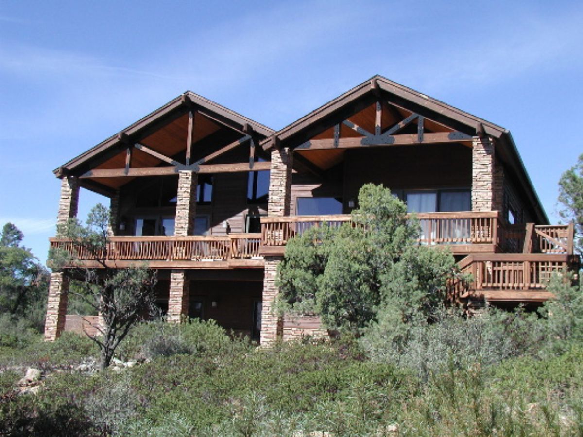 Large rustic mountain cabin with wraparound wooden decks, stone pillars and pine trees under blue sky
