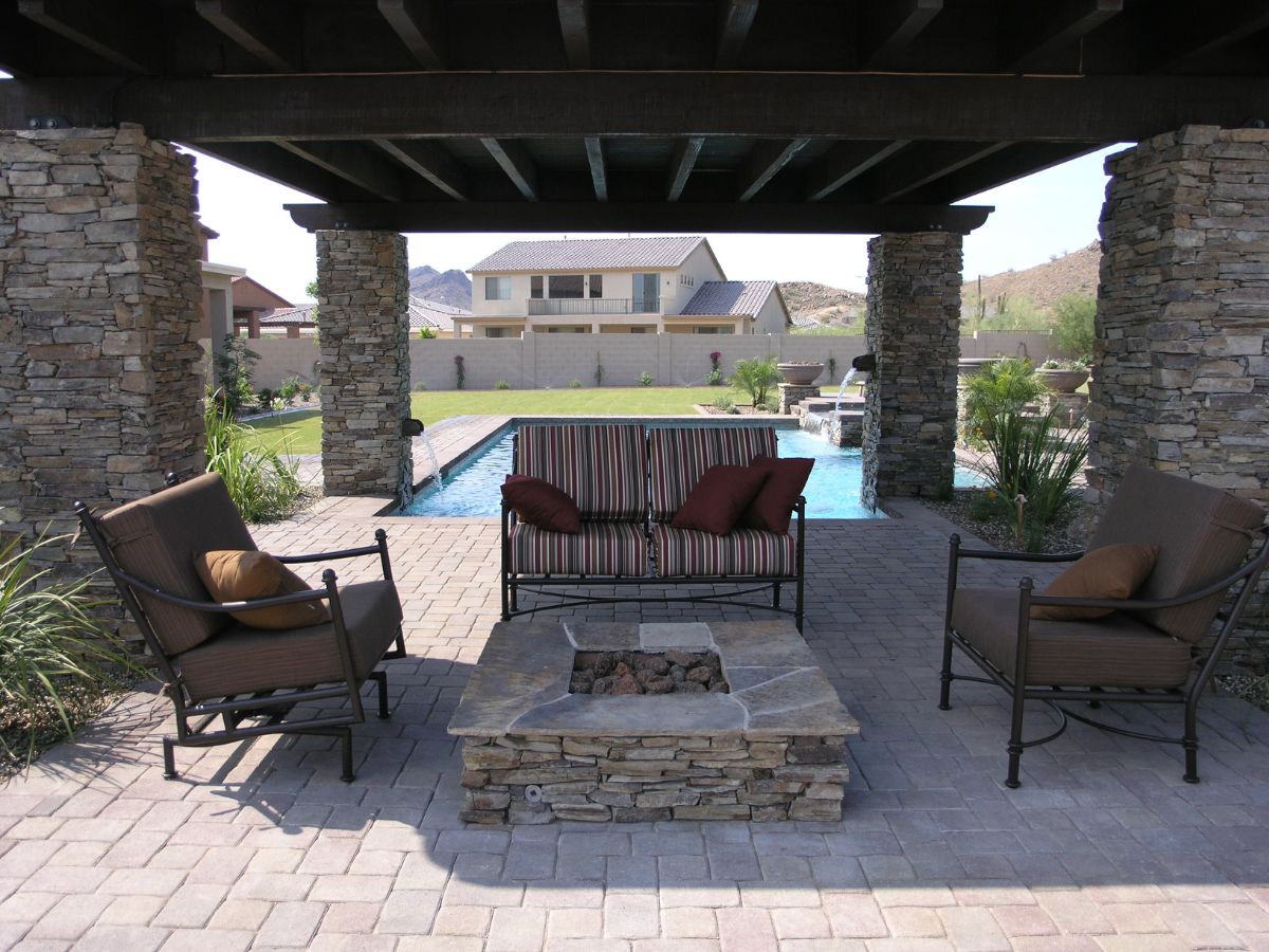 Covered patio with stone pillars, outdoor seating around a stone fire pit, backyard pool and house beyond