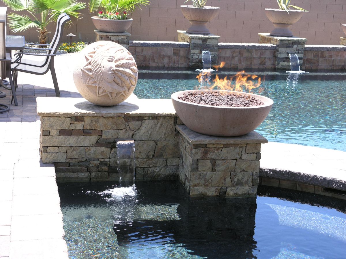 Backyard pool with stone water features, decorative sun sculpture and fire bowl on raised stone ledges by patio chairs