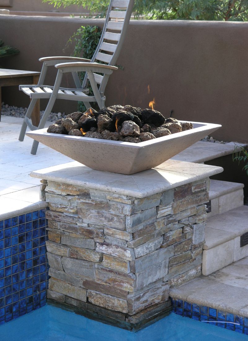 Poolside modern square gas fire pit with lava rocks on stacked stone pedestal beside lounge chair