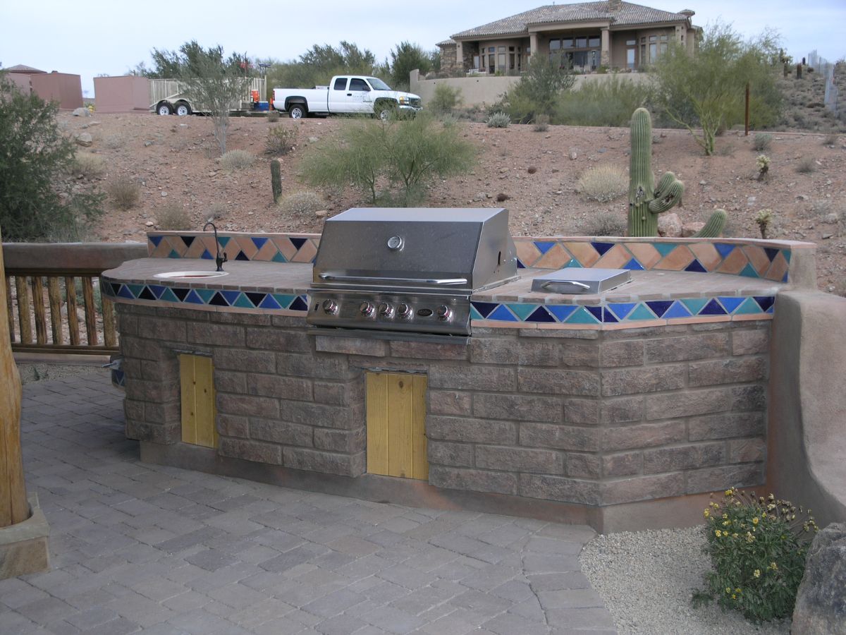 Outdoor kitchen with built-in stainless gas grill, stone base and colorful tile countertop in desert backyard with cactus.