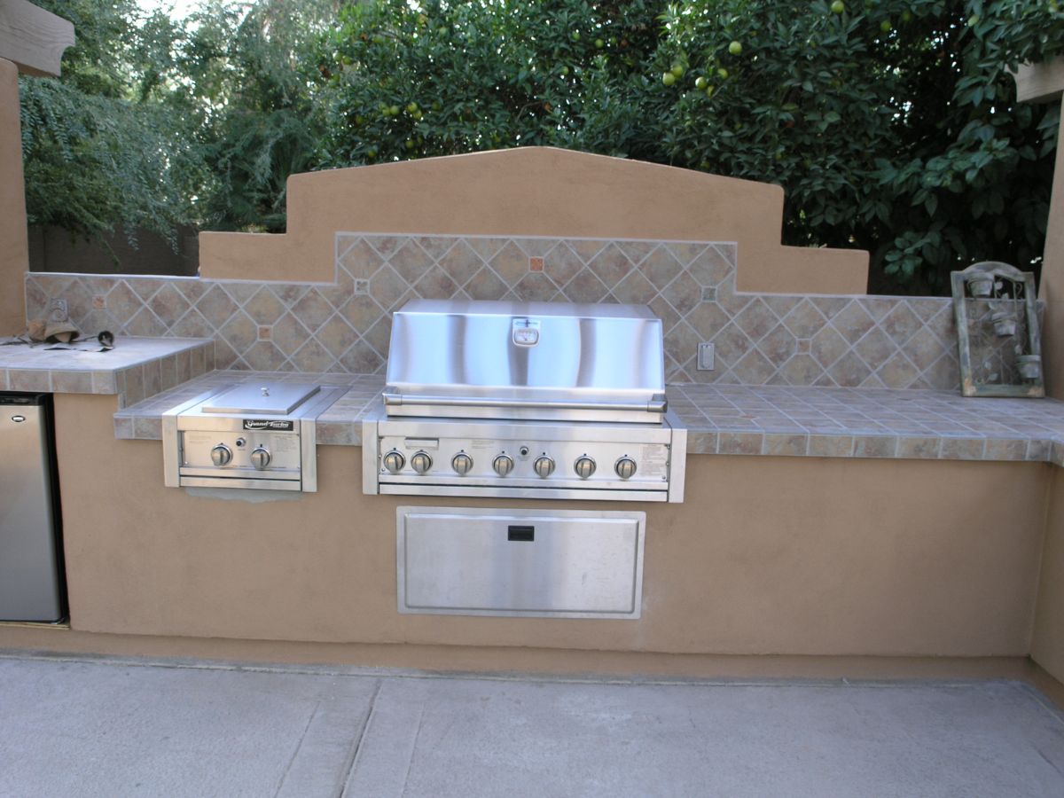 Stainless steel built-in gas grill in tiled outdoor kitchen countertop on a backyard patio with mini fridge and greenery.