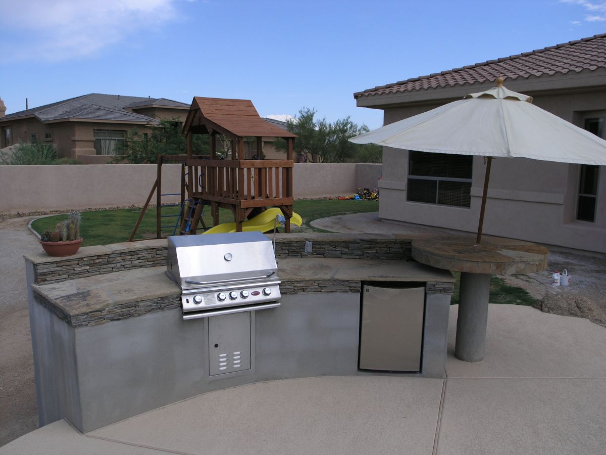 Backyard outdoor kitchen with built-in stainless grill, mini fridge and umbrella table facing a wooden kids' playset.