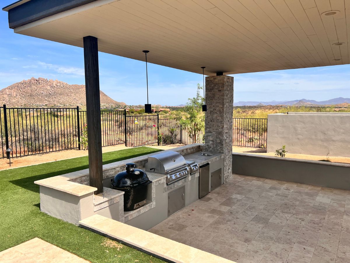 Outdoor kitchen under covered patio with built-in gas grill and kamado smoker, desert mountain view