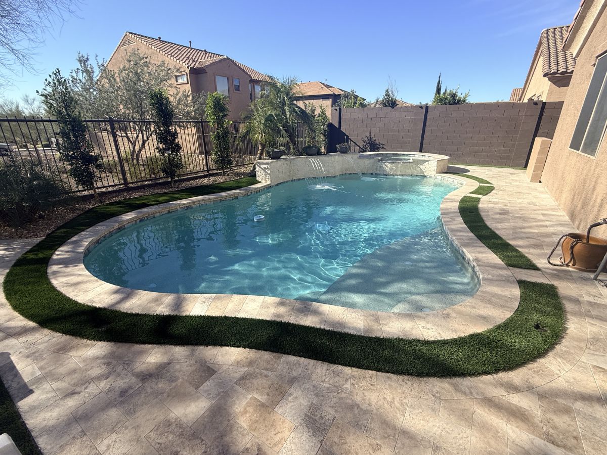 Curved backyard swimming pool with attached spa, travertine paver patio and artificial turf accent under clear blue sky