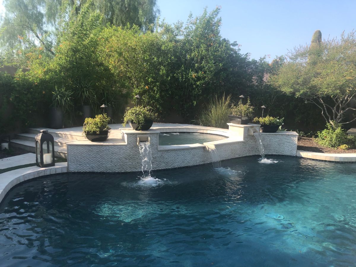 Sunlit backyard pool with raised spa, three cascading waterfalls, potted planters and lush green landscaping