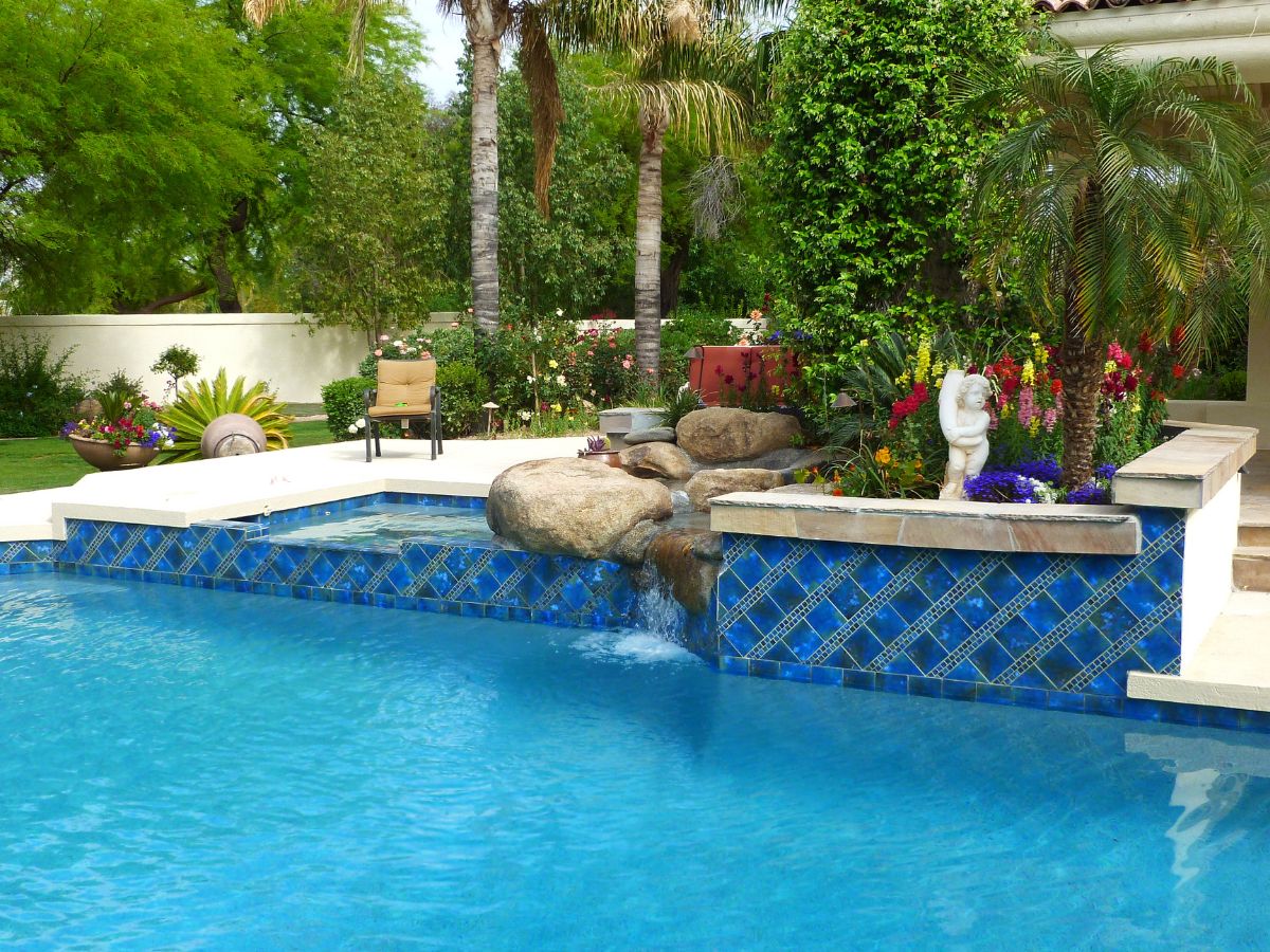 Backyard blue-tiled swimming pool with rock waterfall, spa ledge, tropical palm landscaping and colorful flowering garden.