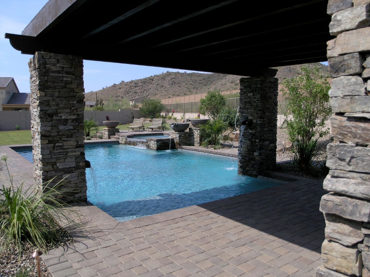Backyard swimming pool with stone columns and pergola, cascading water features, paver patio and lounge chairs in desert yard