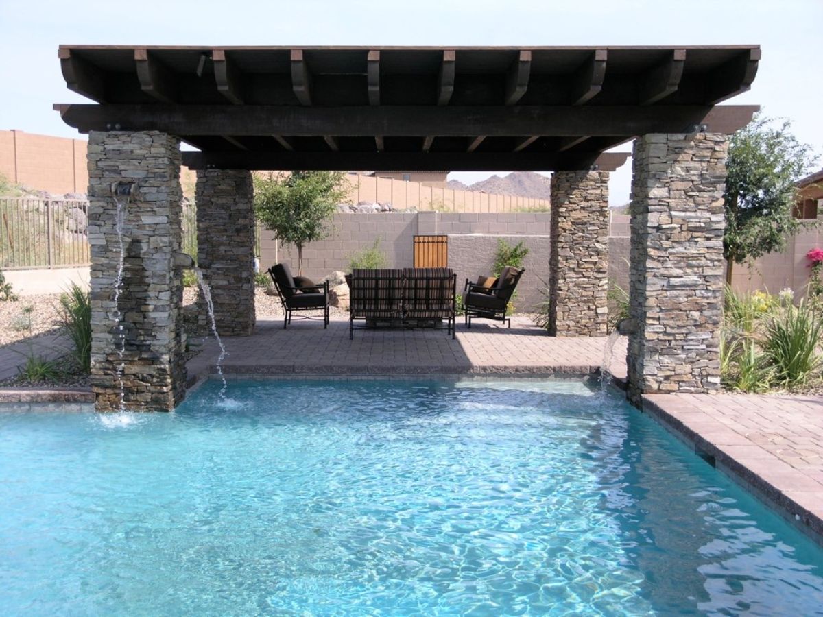 Backyard swimming pool with shaded wooden pergola, stacked-stone columns, cascading water spouts and patio seating