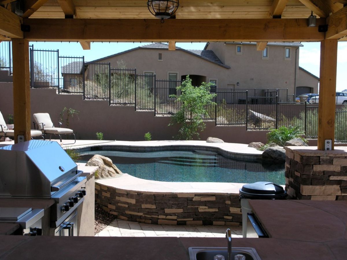 Outdoor backyard patio kitchen with stainless steel grill under wooden pergola overlooking stone-edged pool