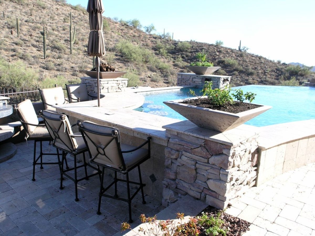 Luxury backyard patio bar with cushioned stools, stone planters and infinity pool overlooking desert hillside
