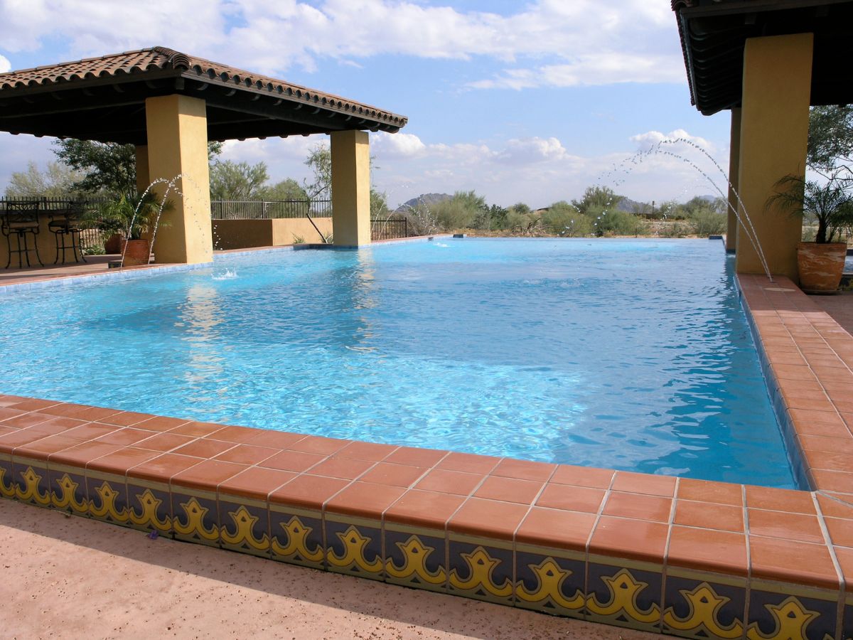 Resort-style outdoor infinity pool with terracotta tile edge, Spanish pavilion and arched water jets in desert landscape