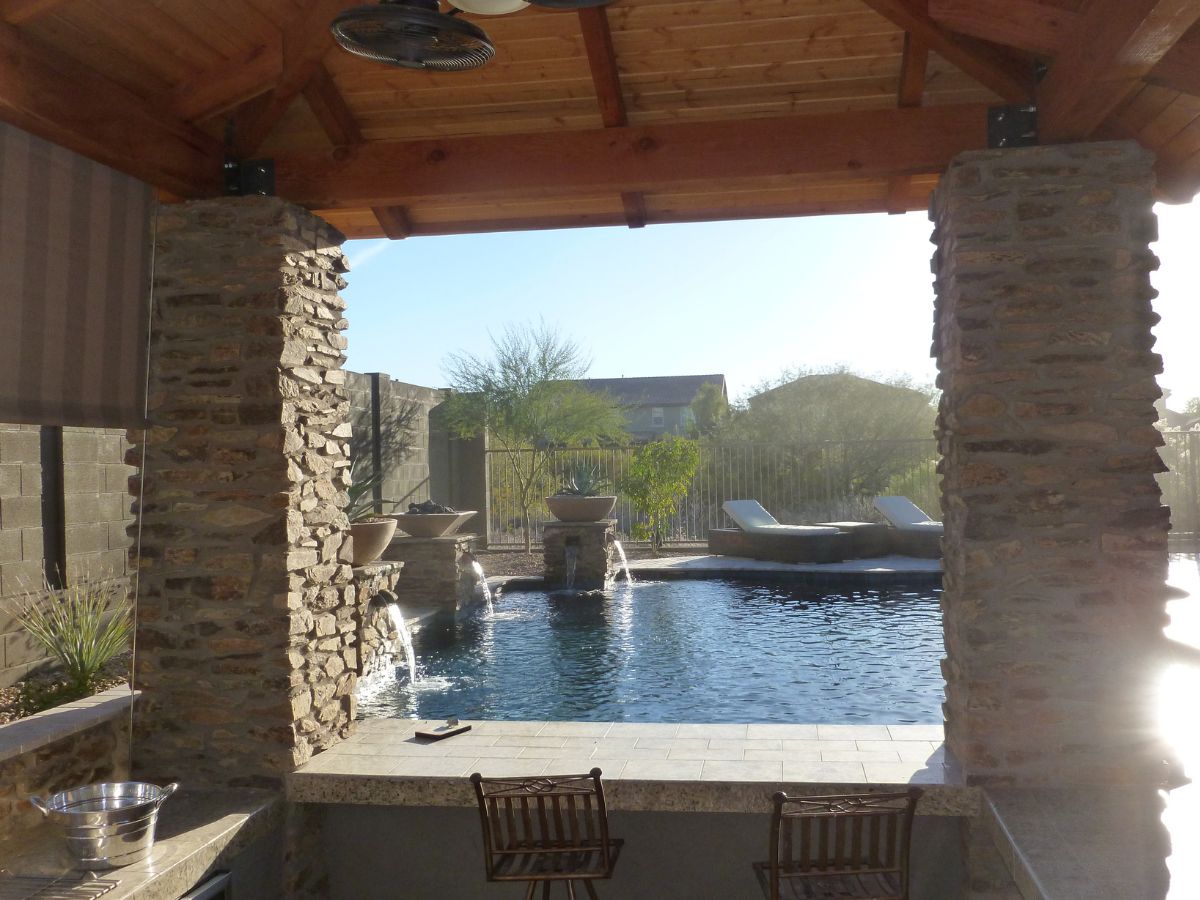 Covered patio with stone columns overlooking backyard swimming pool with cascading water features and lounge chairs