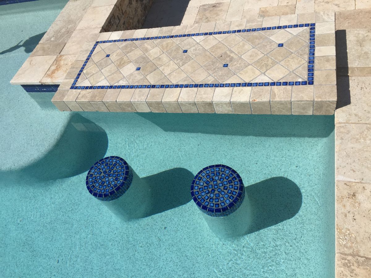 Swim-up bar in pool with two submerged round blue-mosaic bar stools and travertine tiled ledge with blue tile border