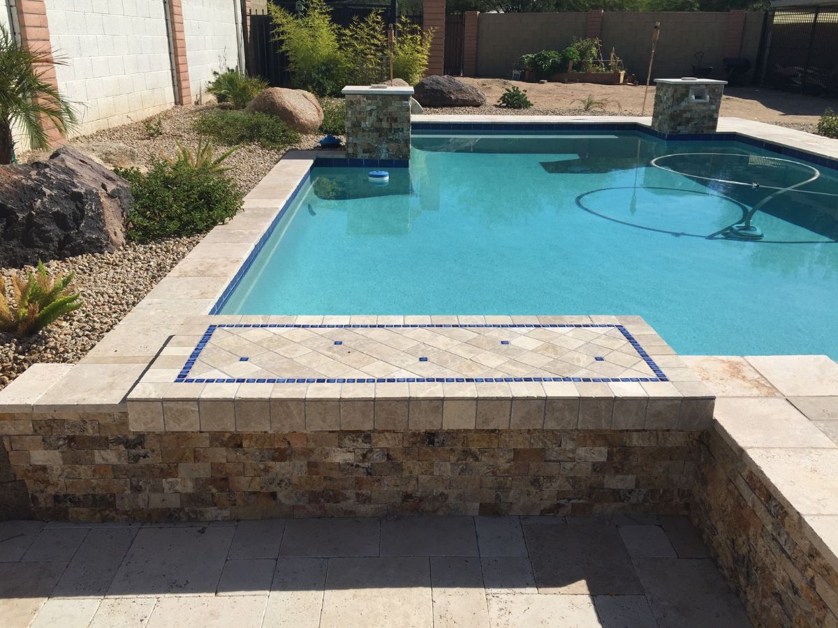 Backyard swimming pool with travertine coping, blue mosaic tile accent and stone veneer wall amid desert rock landscaping