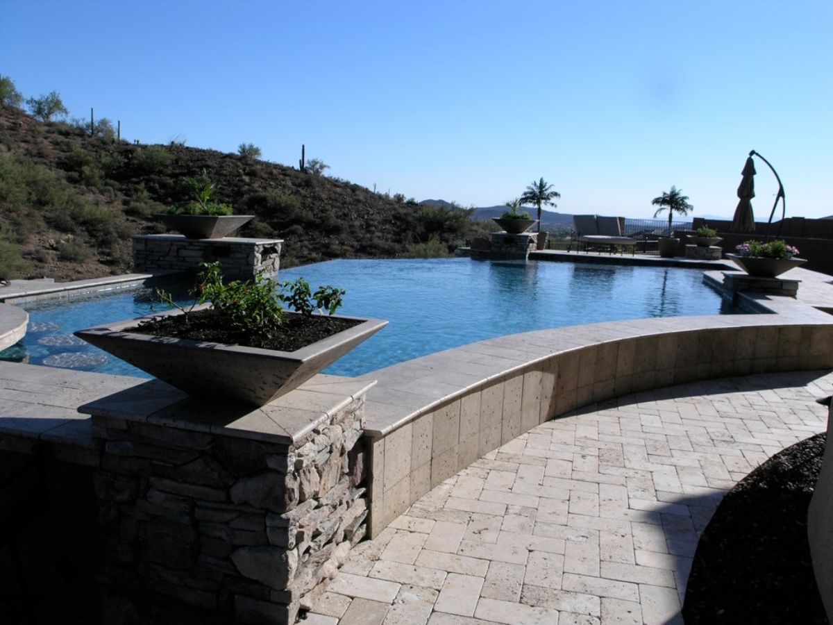 Luxury infinity pool with stone planters, paver patio, lounge seating and desert hillside view under clear blue sky