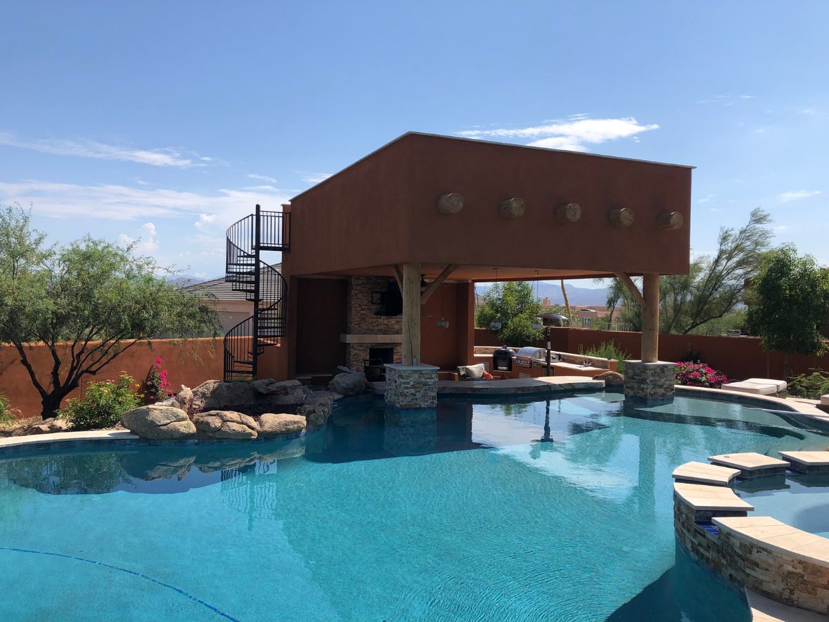 Luxury backyard pool with covered patio and outdoor fireplace, spiral staircase, rock features and desert landscaping.