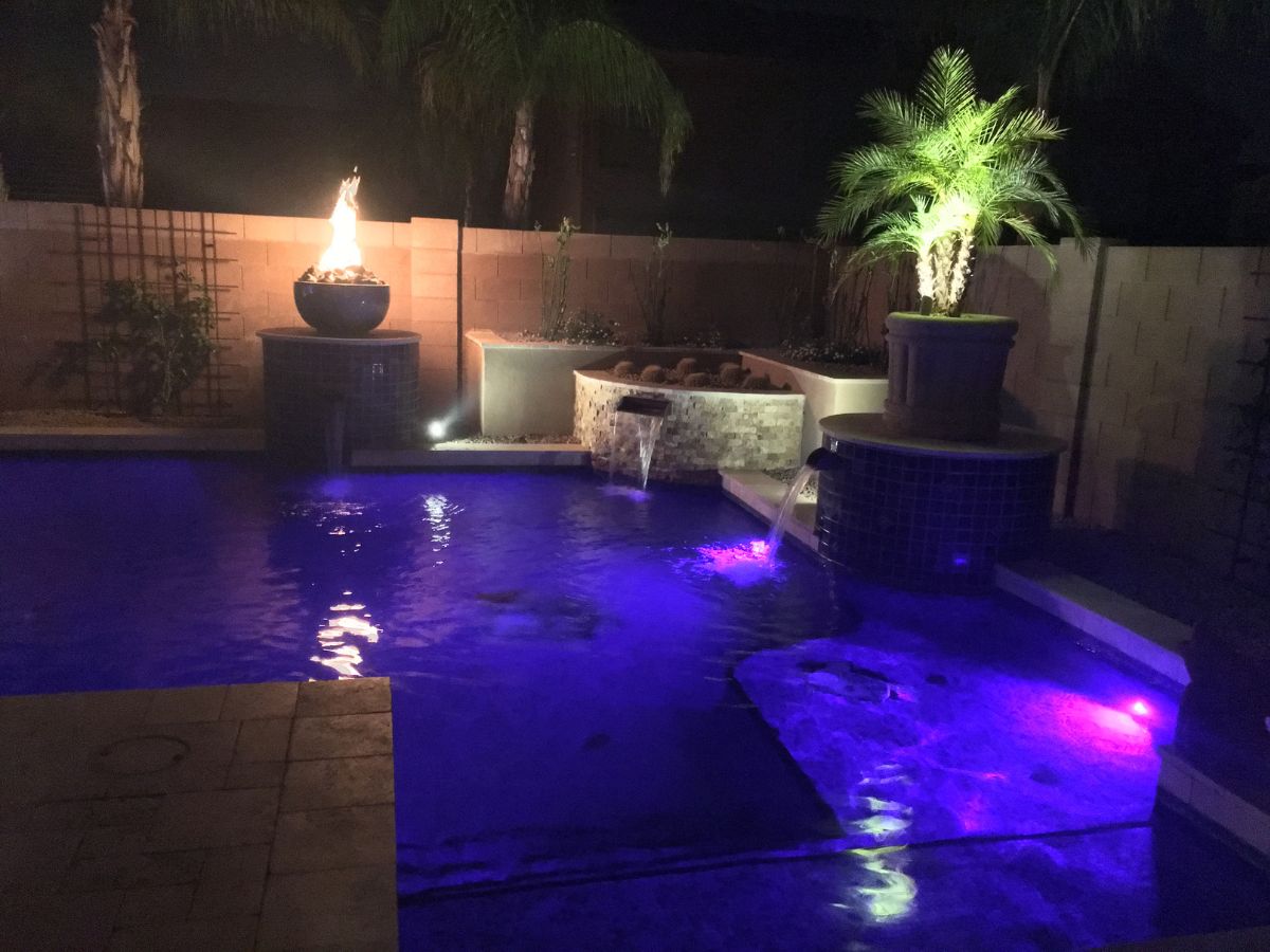 Nighttime luxury backyard pool with purple LED underwater lights, dual water fountains, fire bowl and potted palm