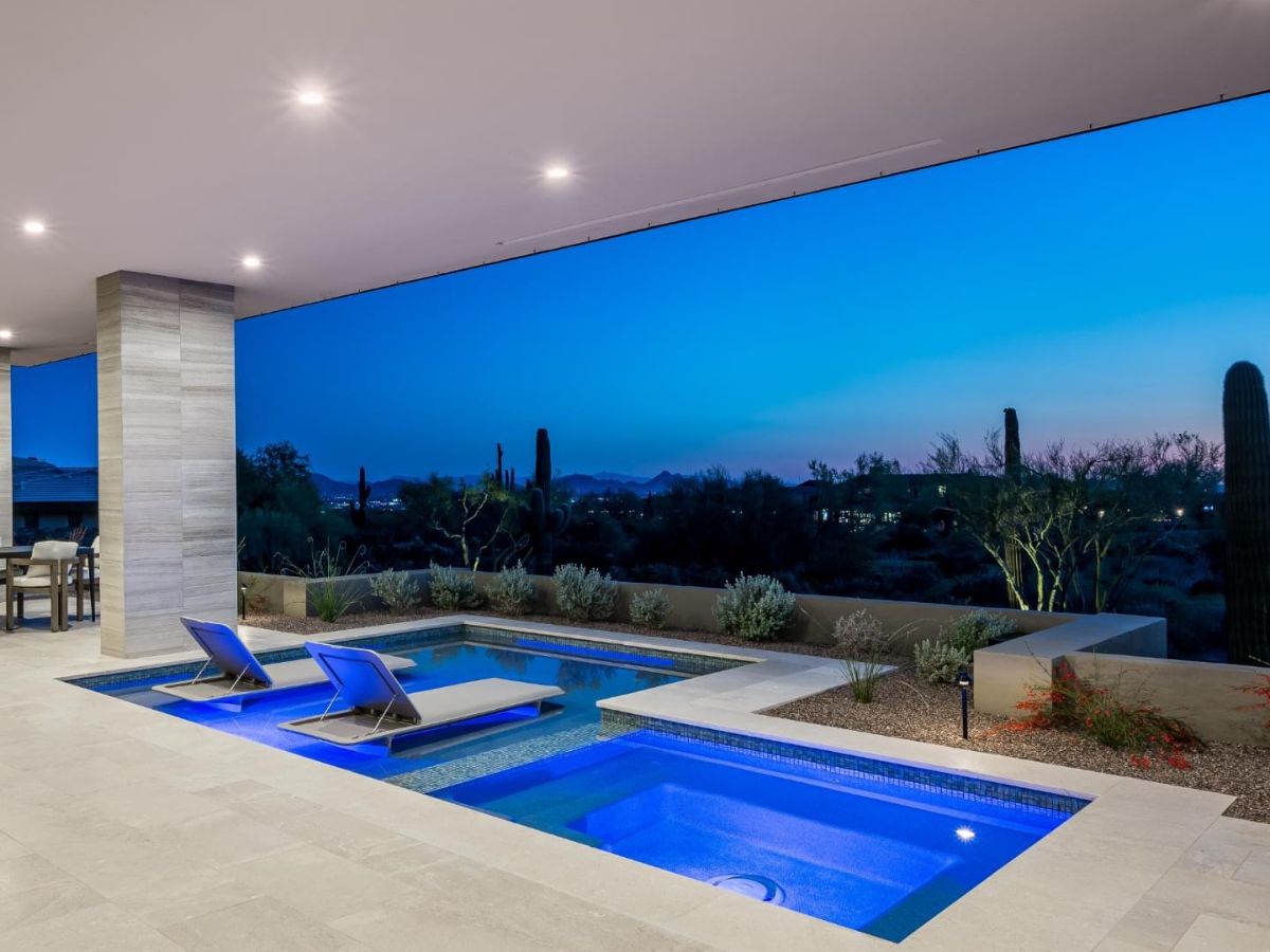 Luxury modern backyard pool with built-in spa, sun loungers on submerged ledge and desert landscape at dusk